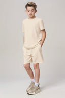 Boy wearing sand beige basic tee and shorts set with beige sneakers, studio background