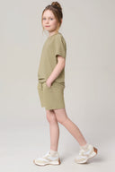 Girl in a light olive green tee and shorts set with white sneakers, standing sideways