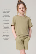 Girl in light olive green cotton t-shirt and matching shorts, hands in pockets, standing indoors.