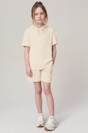 Girl wearing sand-colored hooded tee and shorts set with white sneakers, standing indoors