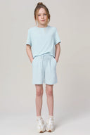 Girl wearing light blue athleisure T-shirt and shorts, hands in pockets, casual studio shot.