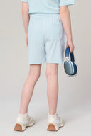Girl wearing light blue athleisure shorts and sneakers holds blue headphones, rear view.