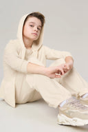 Boy in sand-colored jogger set with hoodie and sneakers, seated against neutral background