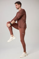 Man in sepia brown athletic tights and long-sleeve top stretching in neutral studio