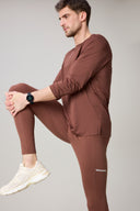 Man stretching in brown long-sleeve activewear and leggings, beige sneakers, neutral background