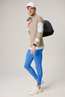 Man in beige athletic jacket and blue leggings carrying black duffel bag, studio background