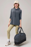 Woman in granite grey long sleeve pima hoodie, yellow leggings, holding gym duffel bag