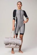 Woman in sleek gray and black rashguard holding beige gym bag with towel, barefoot, athleisure style