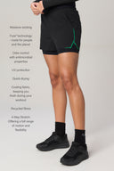 Person wearing charcoal athletic shorts with green accents, showcasing key features like moisture wicking, odor control, UV protection, recycled fibers, and 4-way stretch in a studio setting.