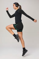 Woman in black long sleeve athletic top and charcoal shorts with green trim, jumping and wearing headphones, activewear studio shot.