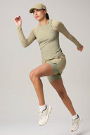 Woman in pale olive athletic shorts, matching long sleeve top and cap, running in studio