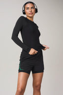 Woman in black long sleeve athletic top and black shorts with green accents, wearing headphones
