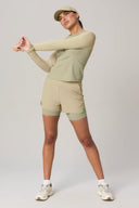Woman in pale olive activewear long sleeve shirt, shorts, and cap stretching in studio.