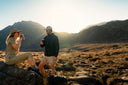 Two people in athleisure wear enjoying drinks outdoors at sunrise in a mountain landscape.